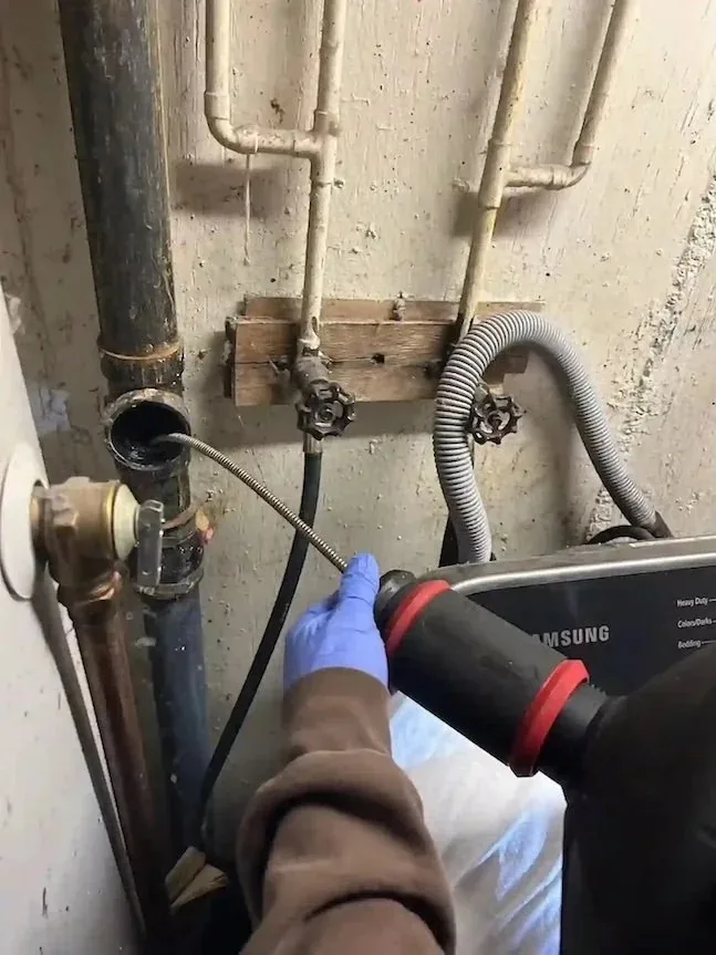 Plumber feeding a motorized auger into a residential drain stack near a washing machine